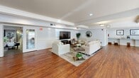 Located at 13430 Coppermine Rd in Herndon, Virginia, this single-floor community space offers a generous and well-appointed layout designed for both gathering and relaxation. The combined dining and living area forms the heart of the space, showcasing rich hardwood flooring, coffered ceiling details, and a classic fireplace that serves as a natural focal point. Multiple dining tables with seating are arranged throughout, complemented by a comfortable lounge area featuring white upholstered sofas, a large flat-screen television, and tasteful décor. A billiard table and upright piano add recreational and cultural character to the room. An abundance of natural light flows in through large windows and glass-paneled doors along the perimeter, offering views of the outdoors and creating a bright, welcoming ambiance. Elegant French doors divide a private meeting or conference area, providing flexible use of the space. The galley-style kitchen is efficiently designed with warm oak cabinetry lining both walls, white appliances including a range, dishwasher, and refrigerator, generous countertop workspace, and a skylight that brightens the room. The kitchen opens conveniently into the main gathering area, making it well-suited for catered events or communal use.
