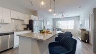 This single-floor residence is designed with a contemporary, open-concept layout that seamlessly connects the kitchen, dining, and living areas. The kitchen is a functional centerpiece, outfitted with white shaker cabinetry, granite countertops, a white subway tile backsplash, and a full suite of stainless steel appliances including a refrigerator, range, microwave, and dishwasher. A large granite-topped island with bar seating serves as both a workspace and casual dining area, illuminated by a cluster of geometric pendant lights. Track lighting runs across the ceiling, adding task-focused brightness throughout the kitchen.

The living area flows naturally from the kitchen and is filled with natural light from oversized windows fitted with horizontal blinds. A ceiling fan adds comfort to the space. The primary bedroom features plush carpeting and connects directly to a private en suite bathroom with a vanity, toilet, and wood-look vinyl flooring. A dedicated walk-in closet area is accessible from the bedroom hallway. A secondary room offers flexibility as a home office or guest space, also carpeted and well-lit by a large window. A second full bathroom with a shower, toilet, and wood-look flooring completes this well-appointed unit.