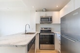 Located along Kingston Road in Toronto's Upper Beaches neighbourhood, this efficiently designed one-bedroom, one-bathroom unit makes excellent use of its single-floor layout. Light wood-tone flooring runs throughout, creating a clean and cohesive aesthetic against the bright white walls and recessed lighting.

The open-concept kitchen and living area serves as the heart of the unit. The kitchen is appointed with sleek white cabinetry, stainless steel appliances including a refrigerator, electric range, and over-the-range microwave, and a striking marble-veined peninsula countertop with an undermount sink — a standout design element that adds both style and functionality. A white tile backsplash completes the polished look.

The bedroom, accessed via a bright hallway, is a cozy private retreat, while the bathroom features neutral tile flooring, a white vanity with a marble-style countertop, a large mirror, and a glass-enclosed shower. The entrance hallway also provides access to a dedicated in-unit laundry area with a stacked washer and dryer — a highly practical inclusion. Natural light filters into the living spaces, enhancing the fresh, modern ambiance of this well-appointed Toronto unit.