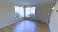 This three-floor space presents a straightforward and functional design with quality finishes throughout. The image showcases a sunlit room featuring light-toned hardwood flooring that extends across the space, lending warmth and a sense of continuity to the interior. Large corner windows dressed with vertical blinds allow generous amounts of natural light to fill the room, creating an airy and open atmosphere. The windows offer pleasant views of surrounding greenery, connecting the interior to the outdoors. The walls are finished in a neutral, warm white tone, providing a clean and versatile backdrop. A floor-level air vent is visible along the baseboard, indicating a built-in HVAC system for climate control. The clean lines, neutral palette, and wood flooring give this level a practical yet inviting character. Spanning three floors in total, the property offers substantial vertical living space, with each level likely providing distinct functional zones. The overall aesthetic is simple and well-maintained, making it a highly adaptable space suitable for a variety of uses and personal styles.