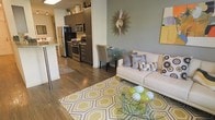 This single-floor residence showcases an open-concept layout that seamlessly integrates the kitchen, dining, and living areas into one cohesive space. The kitchen is equipped with stainless steel appliances, including a gas range, microwave, and refrigerator, complemented by dark cabinetry that extends to the ceiling, providing ample storage. A breakfast bar serves as a natural divider between the kitchen and the adjoining living areas.

Hardwood flooring runs throughout the space, lending warmth and visual continuity. The living area is anchored by a glass-top coffee table and a neutral-toned sofa accented with patterned throw pillows. A bold geometric area rug in yellow and grey tones adds a pop of color and defines the seating zone.

A compact dining area sits adjacent to the living space, furnished with a small table and upholstered chairs, ideal for intimate dining. The accent wall in a muted sage green, paired with a sunburst mirror, adds a decorative focal point. Yellow artwork introduces additional color accents, contributing to the bright and contemporary ambiance of the overall space.