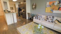 This single-floor residence showcases an open-concept layout that seamlessly integrates the kitchen, dining, and living areas into one cohesive space. The kitchen is equipped with stainless steel appliances, including a gas range, microwave, and refrigerator, complemented by dark cabinetry that extends to the ceiling, providing ample storage. A breakfast bar serves as a natural divider between the kitchen and the adjoining living areas.

Hardwood flooring runs throughout the space, lending warmth and visual continuity. The living area is anchored by a glass-top coffee table and a neutral-toned sofa accented with patterned throw pillows. A bold geometric area rug in yellow and grey tones adds a pop of color and defines the seating zone.

A compact dining area sits adjacent to the living space, furnished with a small table and upholstered chairs, ideal for intimate dining. The accent wall in a muted sage green, paired with a sunburst mirror, adds a decorative focal point. Yellow artwork introduces additional color accents, contributing to the bright and contemporary ambiance of the overall space.