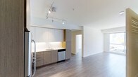 This contemporary single-floor space showcases a thoughtfully designed open-concept layout that seamlessly integrates the kitchen and living areas. The modern kitchen is outfitted with flat-panel cabinetry in a warm taupe finish, a crisp white countertop, an undermount sink with a sleek faucet, and stainless steel appliances including a dishwasher and refrigerator. Under-cabinet lighting adds a polished touch, while a bold yellow accent panel provides a subtle pop of color. Track lighting mounted on the white ceiling ensures flexible, well-distributed illumination throughout the space. The wide-plank hardwood-style flooring in a cool grey tone runs continuously through the kitchen and into the open living area, creating a cohesive and expansive feel. Walls are finished in a clean, bright white, contributing to the airy and light-filled atmosphere. A doorway visible from the main living area leads to an additional room, offering functional separation within the open floor plan. The overall design aesthetic is clean, minimal, and modern, making the space feel both practical and refined.