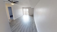 This single-floor residence presents a clean, modern aesthetic defined by neutral gray tones, updated finishes, and consistent wood-look flooring throughout. The open living area is generously proportioned, anchored by a ceiling fan overhead and a textured popcorn ceiling. A sliding glass door leads to a private balcony, allowing natural light to fill the space and providing an outdoor connection. The layout flows naturally from the living area toward the kitchen and bedrooms, as illustrated by the 3D floor plan.

The kitchen features white cabinetry with brushed nickel hardware, speckled gray countertops, and a full suite of black appliances, including a gas range, over-the-range microwave, and refrigerator. The contrast of black appliances against white cabinetry lends a bold, functional character to the space.

The bedrooms are well-sized and bright, each with large windows fitted with vertical blinds that admit abundant natural light. One bedroom includes a full-width mirrored sliding closet, enhancing the sense of space. The bathroom is straightforward and functional, featuring a full tub-shower combination with a curved curtain rod and clean white fixtures. Throughout the unit, crisp white baseboards, textured walls, and updated flooring contribute to a cohesive, move-in-ready atmosphere.