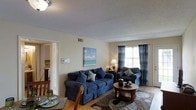 This single-floor residence offers a thoughtfully arranged layout designed for comfortable everyday living. The living room serves as the heart of the home, featuring warm hardwood flooring, a textured ceiling, and a window dressed with plaid curtains that filters in natural light. The space is well-proportioned, providing ample room for relaxed seating arrangements. A coastal-inspired wall painting adds a touch of color and character to the neutral walls, contributing to the space's calm and inviting ambiance. The living area flows naturally into a dining space, visible at the edge of the room, where a dining table and chairs are set for casual meals. A doorway connects to an adjacent room, suggesting an open and accessible floor plan that encourages easy movement throughout the home. The space is finished with practical details including visible HVAC venting and a wall-mounted thermostat, ensuring year-round comfort. Overall, the residence presents a cozy, functional environment with a relaxed coastal undertone, making it a welcoming and livable home.