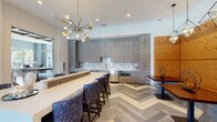 Located at 3585 SW 38th Terrace in Ocala, Florida, this single-floor community clubhouse offers a sophisticated and well-appointed gathering space designed with contemporary flair. Throughout the interior, a distinctive herringbone-patterned tile floor unifies the open layout, complemented by soft blue-gray walls and an abundance of natural light streaming through large windows.

The main lounge area features bold blue marbled upholstered seating, crisp white accent chairs, and a wall-mounted display screen, all beneath a coffered wood-beam ceiling and a striking linear chandelier. Large glass doors open directly to outdoor views of the pool area, enhancing the airy, resort-like atmosphere.

An adjacent game room provides a full-size pool table and additional lounge seating, creating a lively recreational space. The business center is outfitted with built-in workstations, ergonomic seating, and pendant lighting, framed by a large window overlooking lush greenery outside.

The kitchen and bar area is a standout feature, equipped with floor-to-ceiling wood-grain cabinetry, a geometric mosaic tile backsplash, stainless steel appliances, and a long white quartz-topped bar with plush velvet bar stools. An eye-catching Sputnik-style gold chandelier anchors the space with a bold design statement. A cozy banquette dining nook with warm leather upholstering completes the entertaining space.