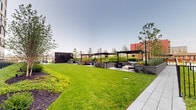 This thoughtfully designed outdoor amenity level offers a comprehensive range of recreational and social spaces within a contemporary urban residential complex. At its centerpiece sits a generously sized lap pool lined with lounge chairs, perfect for relaxation, while a covered cabana area provides shaded retreat nearby. Surrounding the pool, meticulously maintained green lawns and raised planting beds filled with ornamental grasses, shrubs, and young trees create a lush, park-like atmosphere. Multiple distinct seating areas are distributed throughout the deck, furnished with modern sectional sofas and accent chairs in neutral tones with bold red metal frames, encouraging casual gatherings. A games area equipped with a pool table and foosball table adds a lively, social dimension to the space. Sleek light poles and stylish sculptural seating elements enhance the contemporary aesthetic. The deck also features a dedicated terrace with unique pod-style seating and floor-to-ceiling glass walls that reflect the open sky. Panoramic city views, including a prominent suspension bridge on the horizon, frame the entire space, while the surrounding multi-story building facades in light brick and dark metal cladding provide an architecturally striking backdrop throughout.