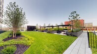This thoughtfully designed outdoor amenity level offers a comprehensive range of recreational and social spaces within a contemporary urban residential complex. At its centerpiece sits a generously sized lap pool lined with lounge chairs, perfect for relaxation, while a covered cabana area provides shaded retreat nearby. Surrounding the pool, meticulously maintained green lawns and raised planting beds filled with ornamental grasses, shrubs, and young trees create a lush, park-like atmosphere. Multiple distinct seating areas are distributed throughout the deck, furnished with modern sectional sofas and accent chairs in neutral tones with bold red metal frames, encouraging casual gatherings. A games area equipped with a pool table and foosball table adds a lively, social dimension to the space. Sleek light poles and stylish sculptural seating elements enhance the contemporary aesthetic. The deck also features a dedicated terrace with unique pod-style seating and floor-to-ceiling glass walls that reflect the open sky. Panoramic city views, including a prominent suspension bridge on the horizon, frame the entire space, while the surrounding multi-story building facades in light brick and dark metal cladding provide an architecturally striking backdrop throughout.