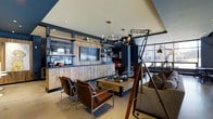 This single-floor space showcases a bold industrial-chic aesthetic that seamlessly blends raw materials with sophisticated design. The main lounge area is anchored by a striking reclaimed wood feature wall housing a flat-screen television and open shelving, displayed against deep navy blue walls that lend the space a moody, refined character.

Seating is thoughtfully arranged to encourage conversation, with a pair of sleek leather and chrome chairs flanking a rustic wooden coffee table on casters, branded "The Local," hinting at a community-focused identity. A plush sofa with patterned cushions adds a softer counterpoint to the room's harder industrial elements.

Overhead, a multi-arm brass and globe chandelier provides warm ambient lighting, complemented by recessed ceiling lights and a dramatic oversized adjustable floor lamp mounted on a tripod stand — a standout design feature that commands attention. A rolling library ladder references classic craftsmanship while adding vertical visual interest.

Large windows along one wall flood the space with natural light and offer views of the surrounding streetscape. Open shelving throughout displays carefully curated decorative objects, vintage finds, and books, reinforcing the space's layered, lived-in personality. The overall ambiance is welcoming yet distinctly stylish — a well-appointed gathering space with strong character and attention to detail.