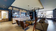This single-floor space showcases a bold industrial-chic aesthetic that seamlessly blends raw materials with sophisticated design. The main lounge area is anchored by a striking reclaimed wood feature wall housing a flat-screen television and open shelving, displayed against deep navy blue walls that lend the space a moody, refined character.

Seating is thoughtfully arranged to encourage conversation, with a pair of sleek leather and chrome chairs flanking a rustic wooden coffee table on casters, branded "The Local," hinting at a community-focused identity. A plush sofa with patterned cushions adds a softer counterpoint to the room's harder industrial elements.

Overhead, a multi-arm brass and globe chandelier provides warm ambient lighting, complemented by recessed ceiling lights and a dramatic oversized adjustable floor lamp mounted on a tripod stand — a standout design feature that commands attention. A rolling library ladder references classic craftsmanship while adding vertical visual interest.

Large windows along one wall flood the space with natural light and offer views of the surrounding streetscape. Open shelving throughout displays carefully curated decorative objects, vintage finds, and books, reinforcing the space's layered, lived-in personality. The overall ambiance is welcoming yet distinctly stylish — a well-appointed gathering space with strong character and attention to detail.