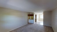 This single-story space features an open-concept layout that connects the living area to the kitchen and an adjoining room. The living area is finished with wall-to-wall carpet in a neutral tone, complemented by smooth, light-colored walls that create a clean and understated aesthetic. A pass-through kitchen counter serves as a natural divider between the living space and the kitchen, offering both visual openness and functional separation. The kitchen features dark wood upper cabinetry and a distinctive patterned backsplash, adding a touch of character to the space. A ceiling-mounted HVAC vent is visible, indicating central air distribution throughout. Beyond the kitchen pass-through, a hallway leads to additional areas of the unit. The adjoining room, visible in the background, benefits from a window that allows natural light to filter in, brightening the space. A ceiling fan is installed in this room, enhancing airflow and comfort. The overall ambiance is calm and neutral, offering a blank canvas well-suited for a variety of furnishing styles.