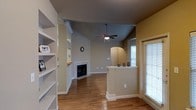 This single-story space is designed with an open-concept layout that promotes a sense of flow and connectivity between living areas. Rich hardwood flooring extends throughout, providing a warm and cohesive foundation underfoot. The main living area draws the eye upward with vaulted ceilings, which are complemented by a ceiling fan with an integrated light fixture, enhancing both comfort and visual appeal.

A classic fireplace with a clean white surround serves as a natural focal point in the living area, adding character and warmth to the space. Built-in shelving lines one wall, offering both display and storage opportunities while reinforcing the room's structured design. A half-wall partition with a flat top surface subtly defines separate functional zones within the open floor plan without compromising the airy feel.

An arched entryway adds an architectural detail that transitions gracefully into an adjacent area, where natural light filters in through blinds on multiple windows. Neutral wall tones throughout the space create a versatile backdrop. The overall ambiance is warm, functional, and well-appointed.