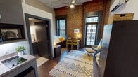 Located at 818 Georgia Ave in the heart of downtown Chattanooga, TN, this urban studio showcases a compelling mix of industrial and contemporary design. The main living area is defined by striking exposed brick walls that rise to impressively tall ceilings, complemented by dark hardwood flooring and a geometric area rug that adds warmth and texture. Exposed ductwork overhead reinforces the industrial aesthetic, while a ceiling fan ensures year-round comfort alongside a wall-mounted mini-split HVAC unit. Large steel-framed windows flood the space with natural light and offer views of the surrounding downtown cityscape. The open layout accommodates a sleeping area with a platform bed and dark wood nightstand, a dedicated sitting nook with a lounge chair and ottoman, and a small dining area with a table and chairs tucked against the brick wall. A compact kitchenette with gray cabinetry and a sink provides everyday functionality. A separate closet area and a full private bathroom with a shower/tub combination, vanity with under-sink storage, and tile flooring complete the unit. The cohesive color palette of charcoal, warm wood tones, and accent yellows creates an inviting, well-curated atmosphere throughout.