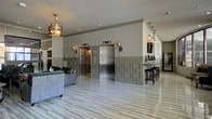 This sophisticated lobby makes a striking first impression with its polished marble tile flooring that reflects light beautifully throughout the space. The interior is defined by a refined two-tone wall treatment, featuring detailed geometric wainscoting in a muted sage-gray finish that adds depth and architectural character to the lower walls, while the upper walls are finished in a soft neutral tone. Two stainless steel elevator doors are centrally positioned, flanked by elegant wall-mounted sconces that provide warm, ambient lighting. A decorative crown-shaped chandelier serves as a bold focal point, adding an element of artistic flair to the lounge area, where a dark leather sofa invites guests to sit and relax. Recessed lighting throughout the ceiling ensures the space is well-lit and bright. To one side, a sleek countertop bar or reception area is lined with dark wood barstools, offering a functional and stylish gathering spot. The overall ambiance is one of understated luxury — polished, contemporary, and thoughtfully designed to balance elegance with comfort, making it an inviting and impressive entry point for residents and visitors alike.