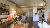 This single-floor residence showcases an open-concept layout that seamlessly connects the living, dining, and kitchen areas into one cohesive and functional space. Rich hardwood flooring flows throughout, anchoring the warm, inviting ambiance created by neutral wall tones and carefully chosen accent colors in coral and orange.

The living area is well-appointed, featuring a plush leather sofa paired with an upholstered accent chair, a glass coffee table, and a patterned area rug that adds visual interest to the space. Decorative elements, including floral arrangements and tasteful artwork, contribute to the polished atmosphere.

Adjacent to the living area, a compact dining space accommodates a round table with seating for several guests, positioned beneath a piece of coastal-inspired wall art. The area benefits from a natural sightline to the front entry, which features a classic paneled door.

The kitchen is positioned along one side of the open floor plan and is distinguished by white upper cabinetry, stone countertops, pendant lighting, and stainless steel appliances. A breakfast bar serves as a natural divider between the kitchen and the main living space, offering additional casual seating and reinforcing the open, connected feel of the interior.