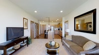 This single-floor residence is designed with an open-concept layout that seamlessly connects the living, dining, and kitchen areas into one cohesive and functional space. The living area is anchored by plush carpeting, a comfortable sofa, and a stylish round coffee table, creating an inviting atmosphere for relaxation. A flat-screen television and framed wall art add to the polished, curated feel of the room.

The kitchen is a standout feature, outfitted with warm honey-toned cabinetry, stainless steel appliances including a full-size refrigerator, and a central island with bar seating. Pendant lighting hangs above the island, providing focused illumination and a modern design accent. Recessed lighting throughout ensures the entire open area is consistently well-lit.

Hardwood-style flooring flows through the kitchen and dining zone, transitioning to soft carpet in the living space, defining each area naturally. Multiple interior doors are visible along the hallway, suggesting several private rooms branching off from the main living space. The neutral color palette of whites and warm beiges, combined with the wood-toned accents, creates a cohesive and welcoming ambiance throughout the residence.