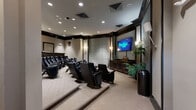 This dedicated movie room is designed to deliver a true theater experience within a sophisticated, well-appointed setting. The space is arranged with multiple rows of plush, black upholstered theater-style seats, complete with built-in cup holders and reclining functionality. The seating is organized on a gently tiered floor plan, ensuring clear sightlines to the large wall-mounted flat-screen display positioned at the front of the room.

The front wall is framed by dark wood millwork and flanked by wall sconces that provide warm, ambient lighting. A substantial wood media console sits beneath the screen, offering storage and housing for audiovisual components. Large acoustic wall panels dominate the side walls, contributing both to the room's sound performance and its clean, modern aesthetic.

The walls are finished in neutral taupe tones, accented by bold dark crown molding and trim that runs throughout, lending the space a polished, cinema-inspired character. Recessed lighting in the ceiling supplements the wall sconces, allowing for adjustable illumination levels. Lush potted plants add a touch of greenery near the front corners.

The rear of the room includes a dedicated entry with a private door, wall-mounted speakers, and a decorative mirror accent, completing the refined, purpose-built design of this private screening space.