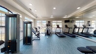 This single-floor community clubhouse is designed with a clean, modern aesthetic and a welcoming coastal-inspired atmosphere throughout. The grand entrance lobby immediately impresses with soaring double-height ceilings, crisp white shiplap walls, elegant crown molding, and expansive grey wood-look flooring that flows seamlessly through the entire space. Decorative columns frame the open corridor leading deeper into the facility, while large windows and recessed lighting ensure the area remains bright and airy.

The lounge area serves as a comfortable gathering space, anchored by a shiplap-accented feature wall housing a sleek fireplace and wall-mounted flat-screen television. Plush charcoal upholstered seating, a rustic wood coffee table, and a woven jute rug create a relaxed, inviting atmosphere complemented by coastal artwork throughout.

The kitchen is outfitted with white shaker cabinetry, subway tile backsplash, dark quartz countertops, and a full suite of stainless steel appliances, including a refrigerator, microwave, and range — making it well-suited for community events and gatherings.

The fitness center is generously equipped with multiple treadmills, elliptical trainers, strength machines, and a weight bench. Large windows allow natural light to fill the room, while a full-length mirror and wall-mounted television complete the well-appointed workout space.