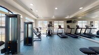 This single-floor community clubhouse is designed with a clean, modern aesthetic and a welcoming coastal-inspired atmosphere throughout. The grand entrance lobby immediately impresses with soaring double-height ceilings, crisp white shiplap walls, elegant crown molding, and expansive grey wood-look flooring that flows seamlessly through the entire space. Decorative columns frame the open corridor leading deeper into the facility, while large windows and recessed lighting ensure the area remains bright and airy.

The lounge area serves as a comfortable gathering space, anchored by a shiplap-accented feature wall housing a sleek fireplace and wall-mounted flat-screen television. Plush charcoal upholstered seating, a rustic wood coffee table, and a woven jute rug create a relaxed, inviting atmosphere complemented by coastal artwork throughout.

The kitchen is outfitted with white shaker cabinetry, subway tile backsplash, dark quartz countertops, and a full suite of stainless steel appliances, including a refrigerator, microwave, and range — making it well-suited for community events and gatherings.

The fitness center is generously equipped with multiple treadmills, elliptical trainers, strength machines, and a weight bench. Large windows allow natural light to fill the room, while a full-length mirror and wall-mounted television complete the well-appointed workout space.