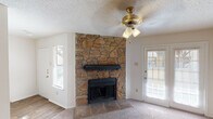 This single-story home is defined by a dramatic floor-to-ceiling natural stone fireplace that anchors the living room, serving as a central architectural focal point. The living area features plush carpeting, a ceiling fan with a brass-toned light fixture, and multiple windows alongside a glass-paned exterior door that allow natural light to fill the space comfortably.

The layout transitions from the carpeted living area into a dining and kitchen zone finished with wood-look hard flooring. The kitchen is configured as a pass-through with a raised breakfast bar, light-toned laminate countertops, white cabinetry, and black appliances. Built-in shelving adjacent to the kitchen adds functional storage and display space. Pendant lighting over the bar area provides warm, focused illumination.

The bedrooms are carpeted and finished with neutral walls and textured ceilings. Each room includes at least one window fitted with blinds, providing natural light and privacy. Closet access is present in the bedrooms.

The bathrooms feature white cabinetry with laminate countertops, large vanity mirrors with overhead lighting strips, and a tub-shower combination with tiled surround. Throughout the home, textured ceilings and white walls create a clean, consistent aesthetic.