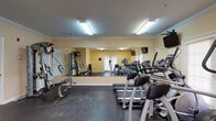 This fitness center is a fully equipped, single-floor facility designed to support a comprehensive workout routine. The space features durable rubber flooring throughout, providing a safe and practical surface for exercise. Crown molding and warm, neutral-toned walls lend a clean, polished aesthetic to the room.

The cardio section is furnished with multiple treadmills and elliptical machines, each positioned in front of a large wall-to-wall mirror that visually expands the space and allows users to monitor their form. Wall-mounted flat-screen televisions are conveniently placed for entertainment during cardio sessions. Natural light filters in through windows with blinds and a set of French doors, creating a bright and welcoming atmosphere.

For strength training, the facility includes a multi-function cable machine suitable for a wide range of resistance exercises, as well as a tiered dumbbell rack stocked with a full range of free weights. Fluorescent overhead lighting ensures the space remains well-lit at all times. The layout is thoughtfully organized to allow users to move freely between equipment. Overall, the fitness center offers a functional and comfortable environment for both cardiovascular and strength-based training.