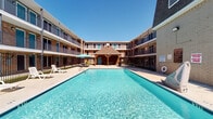 Located at 6425 S Gessner Rd in Houston, Texas, this single-floor residential unit offers a practical and comfortable layout within a multi-building complex. The entryway opens to a tile-floored living area, providing a welcoming transition into the home. Vertical blinds allow natural light to filter into the space, brightening the interior throughout the day.

The kitchen is efficiently designed with dark gray cabinetry offering generous storage, granite-style laminate countertops, a double-basin stainless steel sink, a black electric range, a dishwasher, and a full-size refrigerator. A pass-through opening connects the kitchen to the living area, creating an open and accessible flow between spaces. Adjacent to the living area, a cozy dining nook accommodates a glass-top table with seating for four, accented by a decorative wall clock.

The unit features tile flooring throughout common areas, neutral-toned walls, and recessed panel doors. Residents also enjoy access to the complex's standout outdoor amenity — a large, resort-style swimming pool set within a courtyard framed by the surrounding multi-story brick buildings. A covered gazebo with seating and umbrella-shaded patio furniture complete the outdoor area, ideal for relaxation. The complex is conveniently situated in the Gessner corridor of southwest Houston.
