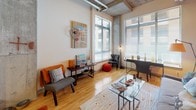 This single-floor urban loft showcases a compelling blend of industrial and contemporary design throughout. The living area immediately impresses with its soaring ceilings, exposed concrete columns, and visible ductwork running overhead — hallmark features of loft-style architecture. Large floor-to-ceiling windows line the space, drawing in abundant natural light and creating an open, airy atmosphere. Light hardwood flooring extends through the main living area, complementing a bold accent wall in a warm orange hue that anchors the room with vibrant character.

The kitchen is well-appointed with dark espresso cabinetry extending to the ceiling for generous storage, stainless steel appliances including a full-size refrigerator, electric range, and microwave, as well as light granite countertops and a stainless steel sink — all set atop the same hardwood flooring.

The bedroom continues the loft's industrial aesthetic with exposed ductwork and concrete architectural elements, softened by plush carpet flooring and a striking deep blue accent wall. Large windows provide ample natural light and outdoor views, enhancing the sense of space.

The bathroom features neutral-toned tile flooring in a linear pattern, a dark vanity with a white countertop, a toilet, and a full-size soaking tub and shower combination with a decorative curtain, completing this well-designed urban retreat.