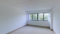 This single-floor space presents a clean, minimalist aesthetic defined by crisp white walls and smooth white ceilings with subtle structural detailing overhead. The interior is finished with wall-to-wall carpeting in a neutral tone, lending a soft and quiet quality to the space underfoot.

A standout feature is the wide bank of black-framed windows that stretch across one wall, flooding the interior with abundant natural light. These windows frame a lush, verdant canopy of mature trees, bringing a sense of the outdoors in and creating a calm, nature-connected ambiance throughout the day.

The layout is open and uncluttered, offering a versatile, functional footprint well-suited to a variety of uses. The restrained, neutral palette throughout — from the pale walls to the light carpet — ensures the space feels airy and adaptable. Overall, the design balances simplicity and comfort, with natural light and greenery serving as the primary design statements.