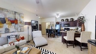 This single-floor residence features an open-concept layout that integrates the living, dining, and kitchen areas into one cohesive, flowing space. The living area is anchored by plush carpeting and benefits from ceiling fan fixtures that help regulate the bright, well-lit atmosphere. A patterned accent rug and chaise lounge chair add character to the lounge area, which transitions effortlessly into the dining space, accommodating a dark wood table and chairs.

The kitchen is a standout feature, showcasing rich, dark espresso-toned cabinetry that extends to the upper walls, providing generous storage space. Black appliances, including a refrigerator, stove, and microwave, complement the cabinetry, while a kitchen island with bar seating creates an additional casual dining or entertaining option. The countertops offer a contrasting lighter tone, adding visual balance to the darker elements.

Throughout the space, the neutral wall tones and light-colored ceilings create an airy, open ambiance. A corridor visible in the background hints at additional private spaces beyond the main living area, contributing to the overall functional and well-organized design of this home.
