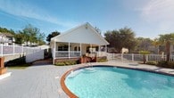 Located at Collins Mill Creek Drive in Milton, Florida, this single-story community space offers a welcoming and functional environment designed for relaxation and gathering. The exterior showcases classic white lap siding beneath a steeply pitched gabled roofline, lending the building a clean, traditional aesthetic that complements its residential surroundings.

A generous covered front porch runs the width of the building, supported by white railings and columns that provide shade and an inviting area to gather. Arched window details add architectural character to the façade, while lush landscaping along the foundation enhances curb appeal.

The centerpiece of the outdoor area is a freeform swimming pool with a brick tile border and a spacious paved deck. The pool area is well-suited for leisure and community activities, with convenient pool entry steps and a broad surrounding deck ideal for lounge seating. White fencing encloses the pool and porch areas, providing both safety and a cohesive visual boundary.

Mature trees frame the property, offering natural shade and privacy. The open sky above and well-maintained hardscape throughout create a bright, airy atmosphere. This space presents a well-appointed community amenity with a charming Southern character, perfectly suited for residents of the Milton, FL area.