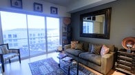 Located in the heart of Dallas, TX, this single-floor residence blends contemporary style with comfortable, thoughtfully curated design. The living area serves as the centerpiece of the space, anchored by a large sectional sofa adorned with patterned accent pillows and grounded by a rich, patterned area rug that adds warmth and texture to the room. A sleek glass coffee table sits at the center, providing a modern focal point.

Floor-to-ceiling sliding glass doors flood the space with abundant natural light and frame sweeping elevated views of the Dallas cityscape, creating a bright and airy atmosphere throughout. The dark accent wall behind the sofa adds depth and contrast, complemented by a large, ornate framed mirror that visually expands the room. Curated wall art and decorative objects reflect an eclectic, well-traveled aesthetic.

A dark etagere bookcase displays curated décor pieces, adding vertical interest and personality to the room. The overall ambiance of the space is sophisticated yet inviting, combining bold design choices with a sense of openness. The elevated vantage point and generous natural light make this Dallas residence a distinctive and memorable space.