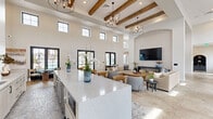 Located at 32449 North Gary Road in San Tan Valley, Arizona, this single-floor community space offers a generously sized, open-plan layout designed for social gathering and leisure. The interior is defined by dramatically soaring ceilings adorned with rustic exposed wood beams and elegant chandelier lighting, creating a grand yet inviting atmosphere throughout.

The living area features a neutral color palette anchored by plush seating arrangements, a substantial wood coffee table, and large-format travertine tile flooring. A statement arched accent wall with a bold geometric tile pattern serves as the focal point, housing a wall-mounted television and a built-in ledge. This same area flows seamlessly into a game room complete with a full-sized billiard table set beneath a contemporary sputnik chandelier.

An expansive bar counter with white quartz surfaces and bar seating lines one wall, accented by dark decorative tile and backed by additional patterned tilework. Clerestory windows and black-framed glass doors line the perimeter, flooding the interior with abundant natural light while providing views of the outdoor amenity areas. The open sightlines and fluid transitions between zones make this space ideal for community events, casual recreation, and relaxed social interaction.