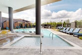 Located at 14020 Stream Way in Charlotte, NC, this thoughtfully designed outdoor amenity space delivers a resort-caliber experience within a modern architectural setting. The centerpiece is a expansive swimming pool with in-water lounge chairs that invite relaxation directly in the water, complemented by a bubbling spa for additional comfort. A series of sleek, wood-slatted pergola cabanas line one side of the pool deck, each outfitted with vibrant teal chaise lounges, offering shaded retreats with an upscale aesthetic. Additional poolside seating with lounge chairs is arranged throughout the open deck area, providing ample space for sunbathing and socializing. A covered indoor-outdoor lounge area anchors one end of the space, featuring soaring ceilings, dramatic floor-to-ceiling glass walls, and bold cylindrical columns. This transitional zone is furnished with cushioned seating and colorful accent chairs around communal tables. A striking brick fireplace with a herringbone tile surround serves as a focal point in the lounge, adding warmth and character. An outdoor kitchen and mounted television complete the entertainment offerings. The surrounding landscaping, open sky views, and cohesive modern design make this a standout communal destination.