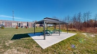 Located at 3859 Sylvan Lakes Blvd in Sylvania, Ohio, this outdoor space offers a generously sized, well-maintained recreational grounds associated with a multi-story brick residential community. The grounds are characterized by expansive open lawn areas ideal for a variety of outdoor activities, bordered by natural wooded edges and seasonal deciduous trees that provide a sense of privacy and connection to nature.

A standout feature of the space is a sturdy, double-tiered metal roof pavilion, anchored on a concrete pad and equipped with multiple green picnic tables. This covered gathering area provides an ideal spot for outdoor dining and community events while offering protection from the elements. The pavilion's durable construction and open-air design make it a functional and welcoming centerpiece of the grounds.

The surrounding landscape includes manicured green lawns with scattered young evergreen plantings and mature trees along the perimeter, creating a layered natural backdrop. Views from the grounds reveal the multi-floor brick residential buildings nearby, along with ample surface parking. The open sky and wide vistas contribute to an airy, expansive atmosphere throughout the outdoor space. Overall, the grounds present a well-appointed, community-oriented outdoor environment suited for relaxation and social gatherings.