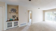 This single-floor residence offers a well-organized layout designed for comfortable everyday living. The spacious living area is finished with plush wall-to-wall carpeting, neutral beige walls, and a textured ceiling, creating a warm and inviting atmosphere. A large sliding glass door dressed with vertical blinds allows abundant natural light to fill the room while providing direct access to the outdoors. A built-in shelving nook adds a practical decorative touch to the living space.

The kitchen is efficiently arranged in an L-shaped configuration, featuring white cabinetry with ample storage, laminate countertops, a double-basin sink, and white appliances including an electric range and dishwasher. Durable wood-look flooring runs throughout the kitchen and bathroom areas, offering easy maintenance.

The bedroom is generously sized with carpeted flooring, a window for natural light, and access to a spacious walk-in closet equipped with multiple hanging rods and shelving — providing excellent storage capacity. An additional door leads to a second closet or utility space.

The bathroom includes a vanity with a white cabinet, a countertop sink, a mirrored medicine cabinet, and wood-look flooring. The overall layout flows logically, connecting each area through wide, accessible doorways for a cohesive and functional living experience.