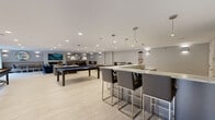 This generously sized single-floor amenity lounge offers a well-appointed, modern space designed for socializing and recreation. The interior is finished with light wood-style flooring throughout, contributing to a bright and airy atmosphere that is further enhanced by recessed lighting and wall-mounted sconces.

The lounge area is elegantly furnished with plush navy blue and taupe armchairs and sofas, arranged around area rugs with colorful accents. Decorative circular mirrors and curated artwork adorn the neutral gray walls, adding visual interest and a contemporary flair. A cozy banquette seating arrangement with a round dining table provides an intimate setting for smaller gatherings.

A dedicated game area features a full-size pool table and a shuffleboard table, both positioned near large windows that flood the space with natural light and offer views of the surrounding greenery. Adjacent to the game area, a sleek bar counter with pendant lighting and bar-height seating is paired with a wall-mounted television, creating an ideal entertainment hub. A pool cue rack nearby underscores the space's recreational focus.

Throughout, the design maintains a cohesive, upscale aesthetic with clean lines, neutral tones, and carefully selected furnishings that balance comfort and style, making this lounge a versatile and inviting communal space.
