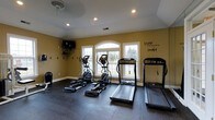 Located at 3900 Suffolk Dr in Dublin, Ohio, this single-floor fitness space is purpose-built for dedicated exercise and wellness. The room is outfitted with rubber-matted flooring, providing both durability and safety underfoot during workouts. A lineup of cardio equipment — including elliptical trainers and a treadmill — is positioned facing a set of elegant arched windows that frame views of the surrounding greenery, allowing natural light to fill the space and creating an open, inviting atmosphere. Recessed ceiling lighting supplements the natural light, ensuring the room is well-lit at any time of day. A wall-mounted television is positioned for easy viewing during exercise, adding a convenient entertainment option. The warm golden-toned walls contribute to an energizing yet comfortable ambiance, while a motivational wall decal reinforces the space's purpose. Hooks along one wall offer practical storage for accessories and equipment. The ceiling fan supports air circulation during intense workouts. Overall, this fitness room is thoughtfully designed to provide a fully functional, comfortable, and motivating environment for a wide range of exercise routines.