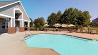 This community amenity space offers a well-rounded combination of indoor and outdoor features designed for comfort and relaxation. The outdoor pool area is a standout highlight, featuring a generously sized swimming pool with a surrounding deck finished in a warm-toned surface. Multiple seating arrangements with chairs and bright green umbrellas provide shaded spots for lounging, while a black iron perimeter fence encloses the area, bordered by lush, mature trees that offer a natural backdrop under open skies.

Inside, the clubhouse is distinguished by soaring vaulted ceilings with striking exposed wood beam accents that lend a warm, rustic-modern character to the space. The main lounge area features rich hardwood-style flooring, a classic fireplace with wainscoting detail, and a wall-mounted television, all complemented by plush tufted velvet sofas and vibrant teal accent chairs arranged around a sculptural wood coffee table. Tall windows flood the interior with abundant natural light, enhancing the airy feel.

A dedicated workspace includes a desk area with workstations, accessible through elegant French doors. The kitchen area is equipped with stainless steel appliances, a center island, and dark cabinetry, while oversized gridded windows frame views of the surrounding greenery, connecting the indoors seamlessly with the natural environment outside.