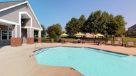 This community amenity space offers a well-rounded combination of indoor and outdoor features designed for comfort and relaxation. The outdoor pool area is a standout highlight, featuring a generously sized swimming pool with a surrounding deck finished in a warm-toned surface. Multiple seating arrangements with chairs and bright green umbrellas provide shaded spots for lounging, while a black iron perimeter fence encloses the area, bordered by lush, mature trees that offer a natural backdrop under open skies.

Inside, the clubhouse is distinguished by soaring vaulted ceilings with striking exposed wood beam accents that lend a warm, rustic-modern character to the space. The main lounge area features rich hardwood-style flooring, a classic fireplace with wainscoting detail, and a wall-mounted television, all complemented by plush tufted velvet sofas and vibrant teal accent chairs arranged around a sculptural wood coffee table. Tall windows flood the interior with abundant natural light, enhancing the airy feel.

A dedicated workspace includes a desk area with workstations, accessible through elegant French doors. The kitchen area is equipped with stainless steel appliances, a center island, and dark cabinetry, while oversized gridded windows frame views of the surrounding greenery, connecting the indoors seamlessly with the natural environment outside.