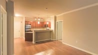 This single-floor space is designed with an open-concept layout that promotes a seamless flow between the kitchen and living areas. The kitchen is a standout feature, showcasing rich cherry-toned wood cabinetry that extends to the upper walls, complemented by stainless steel appliances including a refrigerator and microwave. A peninsula-style breakfast bar serves as both a functional workspace and a natural divider between the kitchen and the adjacent living area, making it ideal for casual dining or entertaining. Pendant lighting hangs elegantly above the bar, while track lighting illuminates the kitchen workspace, ensuring the area is both practical and visually appealing. Crown molding runs along the ceiling throughout, adding a refined architectural detail that elevates the overall aesthetic. The living area is finished with warm hardwood flooring that extends throughout the space, creating a cohesive and inviting atmosphere. The neutral, warm-toned walls allow for versatile interior styling. A white-paneled door provides access to an additional room, suggesting a well-organized and functional floor plan. Overall, the space combines classic design elements with modern finishes to create a comfortable and polished environment.