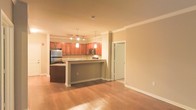 This single-floor space is designed with an open-concept layout that promotes a seamless flow between the kitchen and living areas. The kitchen is a standout feature, showcasing rich cherry-toned wood cabinetry that extends to the upper walls, complemented by stainless steel appliances including a refrigerator and microwave. A peninsula-style breakfast bar serves as both a functional workspace and a natural divider between the kitchen and the adjacent living area, making it ideal for casual dining or entertaining. Pendant lighting hangs elegantly above the bar, while track lighting illuminates the kitchen workspace, ensuring the area is both practical and visually appealing. Crown molding runs along the ceiling throughout, adding a refined architectural detail that elevates the overall aesthetic. The living area is finished with warm hardwood flooring that extends throughout the space, creating a cohesive and inviting atmosphere. The neutral, warm-toned walls allow for versatile interior styling. A white-paneled door provides access to an additional room, suggesting a well-organized and functional floor plan. Overall, the space combines classic design elements with modern finishes to create a comfortable and polished environment.