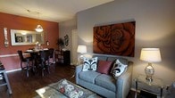 This single-floor space showcases a thoughtfully designed open-concept layout that seamlessly connects the living and dining areas. The living room is anchored by a grey upholstered loveseat adorned with decorative pillows in black, white, and burgundy tones, complemented by a glass-top coffee table. A striking large-scale two-panel artwork depicting roses in rich, deep browns serves as a dramatic focal point on the grey accent wall, adding depth and artistic character to the room.

Dark hardwood flooring runs throughout, providing a warm and grounding contrast to the neutral grey walls and the bold burnt-orange accent wall that defines the dining area. The dining space features dark wood furniture and seating, with a pendant light fixture overhead that adds ambient warmth. A sideboard with decorative storage drawers sits against the wall, topped with plants and decorative accessories. A mirror on the dining room wall further enhances the sense of openness. The overall ambiance is cozy yet sophisticated, with a rich, earthy color palette that creates an inviting and stylish atmosphere throughout.