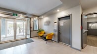 Located at 235 Willis Cres in Saskatoon, Saskatchewan, this building lobby offers a polished and functional common space designed to make a strong first impression. The entrance hallway greets visitors with large glass double doors that flood the space with natural light, large-format light-toned ceramic tile flooring, and recessed ceiling lighting that ensures a bright and welcoming atmosphere. A grey entrance mat anchors the entryway, while a pair of bold yellow accent chairs and a small side table provide a casual seating nook for residents and guests alike.

The adjacent living room lounge extends the space with a more relaxed, residential feel. A plush grey sectional sofa and chaise are complemented by a decorative round metal mirror and a large-scale abstract canvas artwork in muted blue and gold tones, lending an air of sophistication. Potted greenery adds life and warmth throughout.

A standout feature of the lounge is a sleek, modern linear fireplace set within a textured stacked-stone surround, serving as a dramatic focal point on the main wall. A framed bulletin board provides practical resident information, rounding out the functional aspects of the space. Together, the hallway and lounge create a cohesive, contemporary lobby environment that balances comfort with modern design.