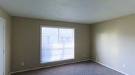 This single-floor residential space offers a clean and functional layout designed for comfortable everyday living. The interior is finished with warm, neutral-toned walls that create a calm and cohesive atmosphere throughout. The flooring is covered in plush carpeting, adding softness underfoot and contributing to a cozy, quiet environment.

A notable feature of the space is the large picture window fitted with horizontal blinds, allowing for generous amounts of natural light to fill the room while maintaining privacy control. The window provides a view of the surrounding exterior, bringing a sense of openness to the interior. The textured popcorn ceiling adds a classic residential character to the space and runs consistently throughout.

White baseboards provide a crisp contrast against the taupe wall color, giving the room a tidy, well-defined finish. The overall design is straightforward and practical, with a neutral palette that serves as a versatile backdrop for a variety of furnishing styles. The single-floor layout ensures ease of movement and accessibility, making the space well-suited for a range of occupants. The combination of natural light, soft flooring, and understated finishes creates an inviting and livable environment.