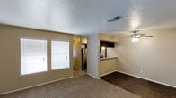 This single-story Garland property offers an open and functional layout that integrates the living, dining, and kitchen areas into a cohesive shared space. The living area is finished with plush carpeting, while the adjoining dining and kitchen zones transition to hardwood-style flooring, creating a natural visual distinction between spaces. A ceiling fan with a light fixture serves the dining area, providing both airflow and ambient lighting. Natural light enters through a set of windows fitted with horizontal blinds, brightening the main living space. The kitchen features a raised pass-through counter with a granite-style surface, dark cabinetry, and stainless steel appliances, including a refrigerator visible from the main living area. The pass-through design enhances the open feel and allows easy interaction between the kitchen and the rest of the living space. Walls are finished in a warm neutral tone throughout, complemented by a textured ceiling. A hallway visible from the main area leads to additional rooms, suggesting a well-organized floor plan. The overall ambiance is practical and comfortable, suitable for everyday living.
