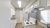 This single-floor space includes a well-appointed kitchen designed for practicality and efficiency. The kitchen features white cabinetry that lines the upper walls, providing ample storage, complemented by a range hood positioned above a white electric coil-burner stove. A full-sized white refrigerator anchors one end of the kitchen, while a double-basin sink with a standard faucet sits along the opposite counter, offering a functional layout for meal preparation. The countertops display a speckled, neutral-toned finish, consistent throughout the kitchen workspace. Light wood-look flooring runs through the space, adding warmth and contrast to the predominantly white cabinetry and appliances. Overhead, a flush-mounted fluorescent light fixture provides bright, even illumination across the kitchen. The walls are finished in a neutral gray tone, and the textured ceiling adds subtle visual interest. A door visible at the end of the kitchen leads to an adjacent area, suggesting a connected layout within the unit. Overall, the kitchen presents a clean, cohesive aesthetic suited for everyday use.