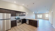 This single-floor home offers a thoughtfully designed open-concept layout that seamlessly connects the kitchen, dining, and living areas. The kitchen is a standout feature, showcasing rich dark cabinetry, granite countertops, a peninsula with a dual-basin sink, and a full suite of stainless steel appliances including a range, microwave, dishwasher, and refrigerator. Elegant pendant lighting and recessed fixtures illuminate the space, while soaring vaulted ceilings lend an impressive sense of openness and volume. Durable wood-look plank flooring runs throughout the main living areas, providing a warm and cohesive aesthetic. The living area benefits from natural light streaming through large windows that frame views of the surrounding outdoor greenery, with a ceiling fan adding comfort and character. The bedrooms are carpeted for comfort and feature ceiling fans, generous windows, and walk-in closets with built-in shelving. The primary suite connects directly to a well-appointed en-suite bathroom complete with dark cabinetry, granite countertops, and a walk-in closet with multi-tier shelving. The entry foyer welcomes visitors with natural light and direct views of the landscaped exterior. Throughout the home, neutral warm-toned walls and white trim create a clean, move-in-ready canvas.