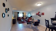 This single-floor residence makes a strong visual statement through its cohesive red, black, and white color scheme carried throughout the space. The open-plan living and dining area is anchored by rich burgundy leather seating arranged around a glass-top coffee table atop a striped area rug. Natural light filters through dark-curtained windows, complementing the cool grey walls and light tile flooring. A glass dining table with sleek black chairs sits adjacent to the living area, accented by bold red abstract wall art, creating a seamless flow between entertaining zones. The bedroom is a standout feature, boasting a dramatic floor-to-ceiling striped accent wall in red and black that lends a bold, design-forward character to the room. A queen bed with a patterned rug and a window with decorative curtains complete the space. The kitchen offers ample warm-toned wood cabinetry, generous countertop space, a double stainless steel sink, and under-cabinet lighting for practical functionality. The bathroom is finished with tile flooring, a full tub-shower combination, and a wood-toned vanity. Consistent tile flooring and textured ceilings unify the home's interior throughout.
