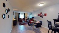 This single-floor residence makes a strong visual statement through its cohesive red, black, and white color scheme carried throughout the space. The open-plan living and dining area is anchored by rich burgundy leather seating arranged around a glass-top coffee table atop a striped area rug. Natural light filters through dark-curtained windows, complementing the cool grey walls and light tile flooring. A glass dining table with sleek black chairs sits adjacent to the living area, accented by bold red abstract wall art, creating a seamless flow between entertaining zones. The bedroom is a standout feature, boasting a dramatic floor-to-ceiling striped accent wall in red and black that lends a bold, design-forward character to the room. A queen bed with a patterned rug and a window with decorative curtains complete the space. The kitchen offers ample warm-toned wood cabinetry, generous countertop space, a double stainless steel sink, and under-cabinet lighting for practical functionality. The bathroom is finished with tile flooring, a full tub-shower combination, and a wood-toned vanity. Consistent tile flooring and textured ceilings unify the home's interior throughout.