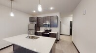 This single-floor unit is designed with a clean, contemporary aesthetic that emphasizes open living and functional design. The open-concept layout connects the kitchen and living area seamlessly, creating a spacious and airy atmosphere ideal for both everyday living and entertaining.

The kitchen is a standout feature, showcasing rich dark-stained cabinetry that extends to the ceiling for ample storage, paired with crisp white quartz-style countertops that provide a striking contrast. A central island with an undermount sink and pendant lighting above adds both practicality and style. The kitchen is fully equipped with a full suite of stainless steel appliances, including a refrigerator, electric range, and over-the-range microwave.

The living area is generously proportioned with light wood-style plank flooring that flows consistently throughout the unit, contributing to a cohesive and modern feel. Neutral white walls and recessed lighting keep the space bright and versatile.

The bathroom continues the unit's contemporary design language, featuring dark wood-toned vanity cabinetry, a white countertop with an integrated sink, a large vanity mirror, and light flooring that complements the rest of the unit. The layout is efficient and well-appointed.

Overall, this unit offers a well-balanced combination of modern finishes, thoughtful storage solutions, and an open floor plan that maximizes the sense of space.